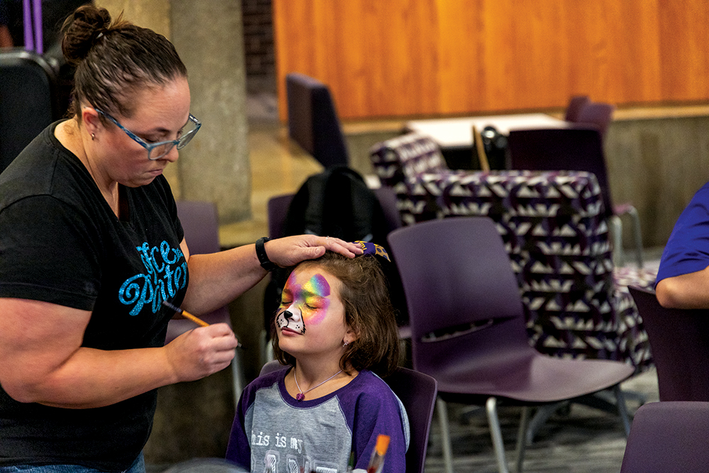 Young girl getting her face painted by a woman in a black shirt.
