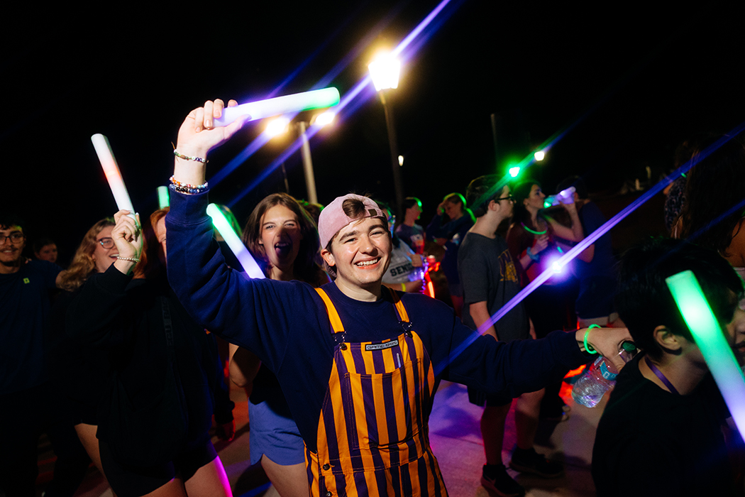 Student on campus at night in a purple sweatshirt and yellow and purple striped overalls with their hands up holding a glow stick.